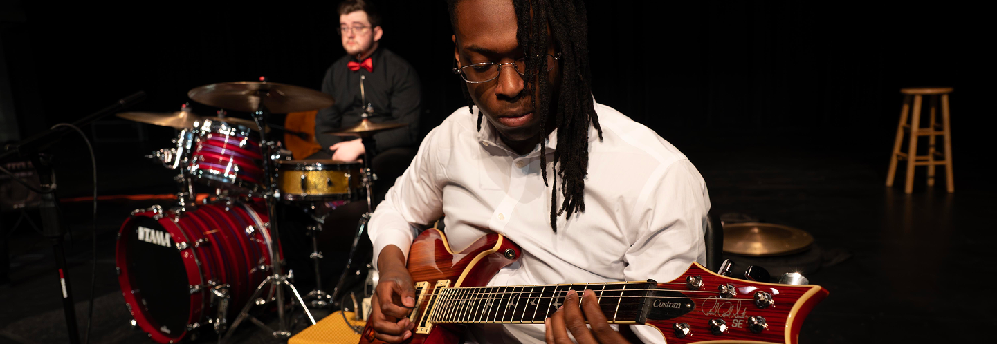 Close-up of student playing guitar during Jazz Combos performance. Student playing drumset in the background.
